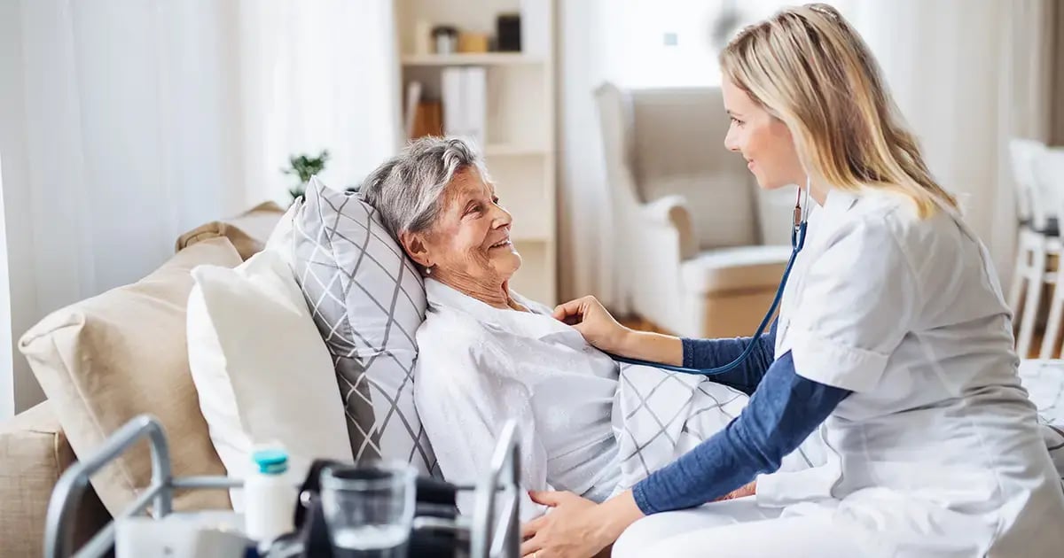 An elderly woman receiving a home medical examination from a female doctor. Both are smiling.