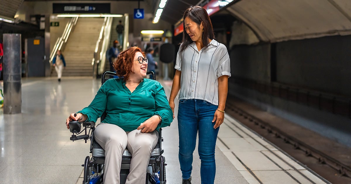 Frau mit Multipler Sklerose und Assistenzperson an einer Bahnhofsstation