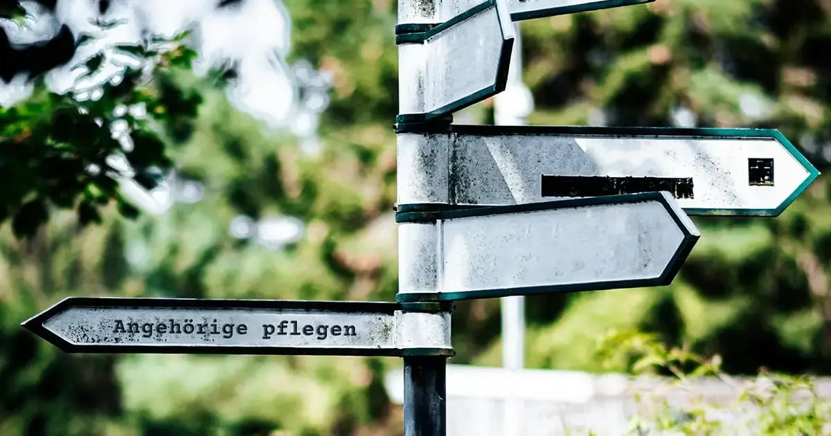 Image of street signs. On the one pointing in a different direction from the others, the German text reads “Angehörige pflegen” (“Caring for relatives”).
