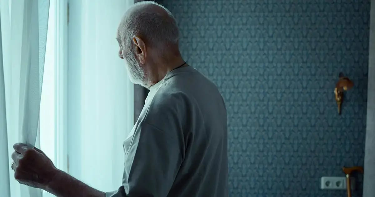 Depressiver, älterer Mann in dunklem Zimmer schaut aus dem Fenster, durch das schwach Tageslicht dringt.