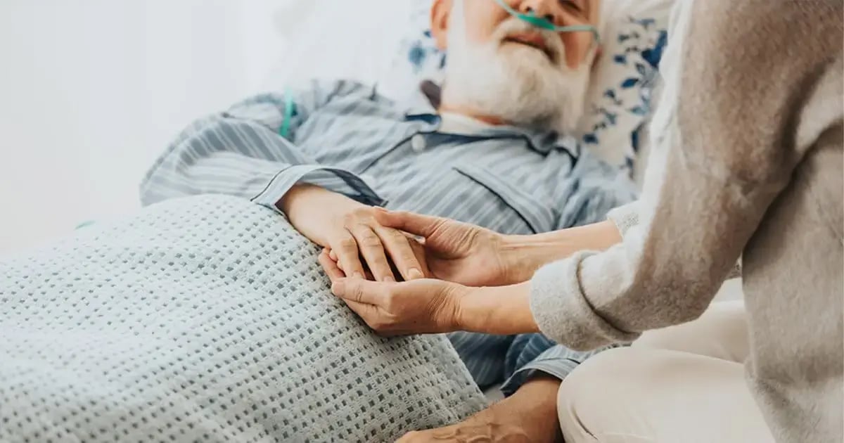 Pflegende Angehörige hält sanft die Hand eines älteren Patienten im Bett, fürsorgliche Palliativpflege, Fokus auf den Händen, die Geborgenheit vermitteln.