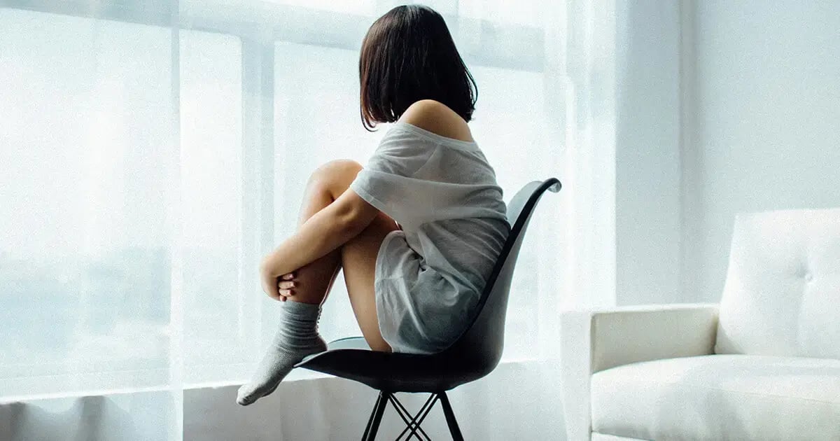 Woman sitting on a chair, hugging her knees, gazing out the window — symbolic image for depression.