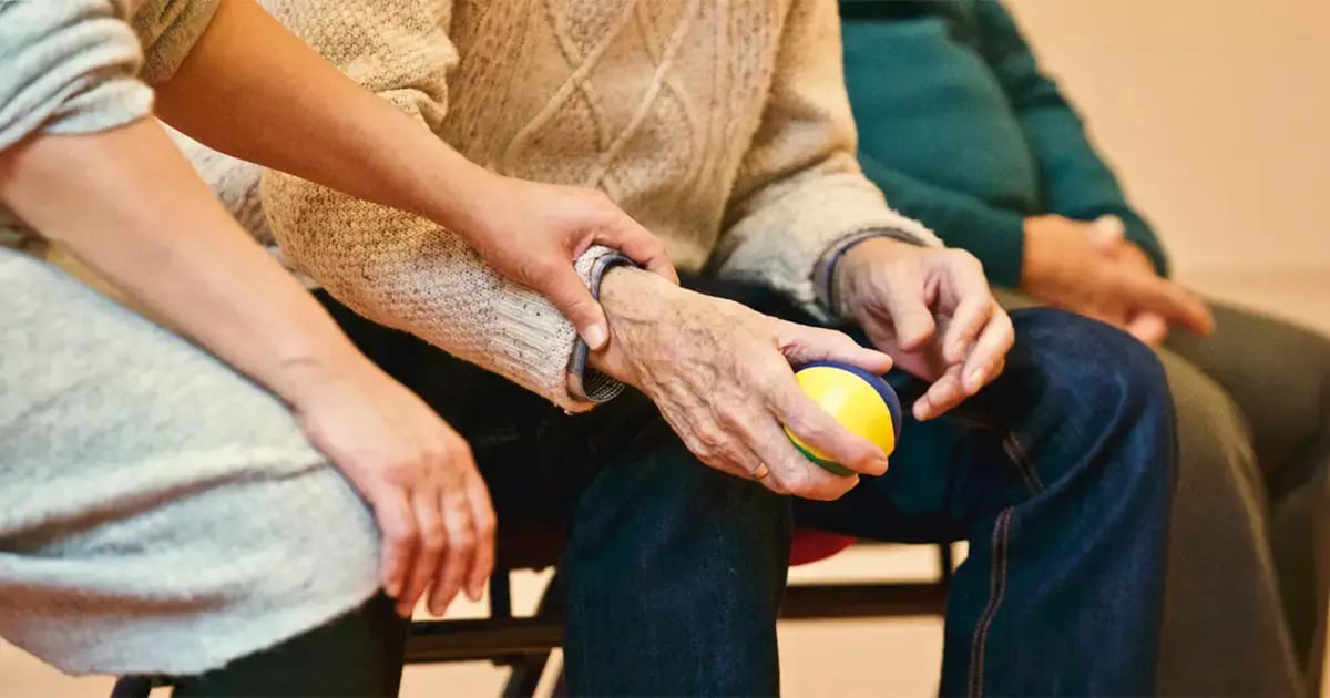 2 Personen sitzen nebeneinander. An den Händen lässt sich erkennen, dass eine vermutlich alt, die andere jung ist. Alte Hand hält einen Ball